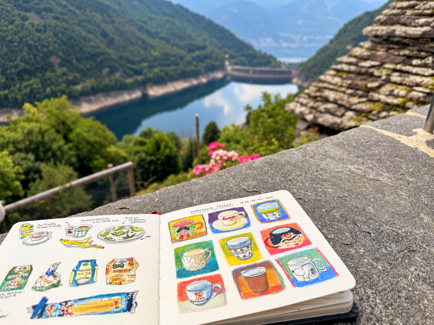 Aufgeschlagenes Skizzenbuch auf einer Terrasse mit Blick auf den See und die Berge.